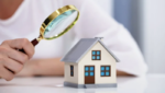A person with a magnifying glass looking at a miniature home simbolyzing property title search